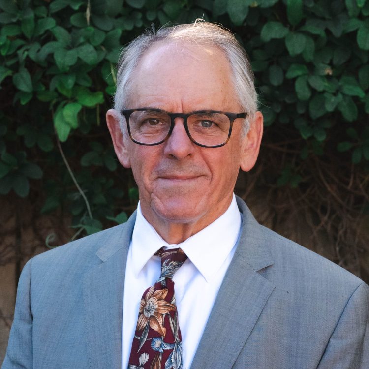 Richard Davies. A white man with white hair. He is wearing glasses. He is wearing a grey blazer over a white shirt with a red, floral tie.