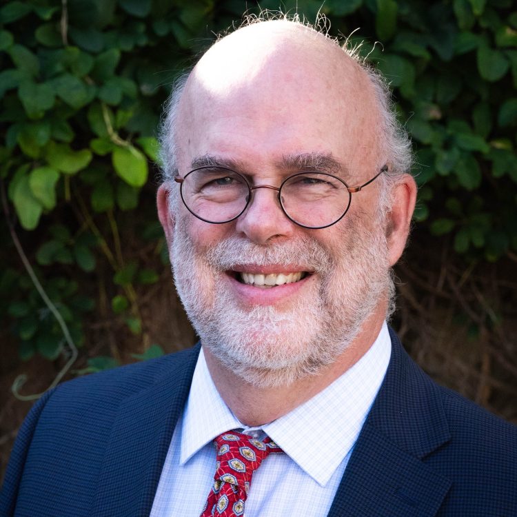 Scott Charlton. A white man with white hair. He is wearing glasses. He is wearing a blue blazer over a white checked shirt with a red tie.